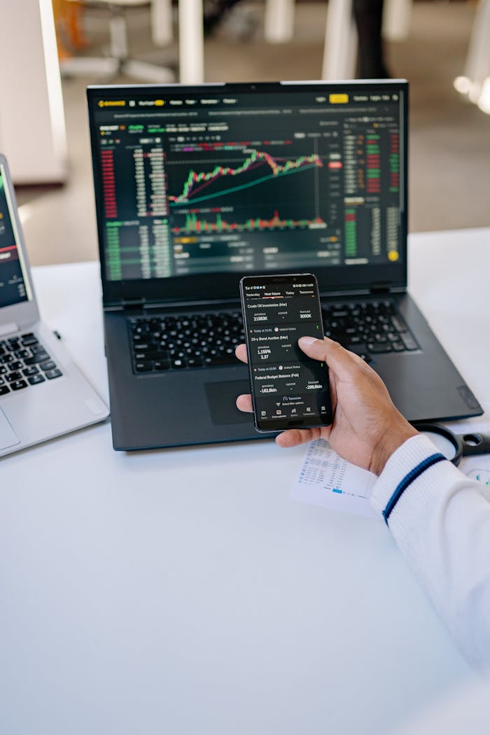 Person analyzing stock market data on a laptop and smartphone at a desk.